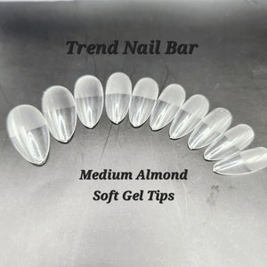 May include: Clear, medium almond-shaped soft gel nail tips. The image shows a set of clear, oval-shaped nail tips, arranged in a semi-circle. The text "Trend Nail Bar" and "Medium Almond Soft Gel Tips" are visible.