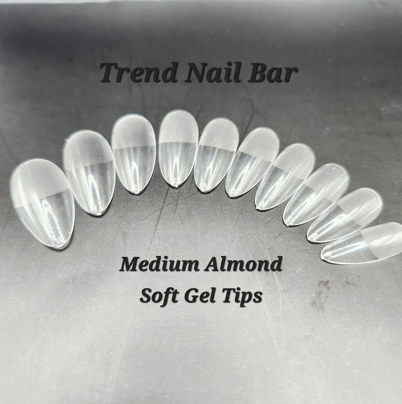 May include: Clear, medium almond-shaped soft gel nail tips. The image shows a set of clear, oval-shaped nail tips, arranged in a semi-circle. The text "Trend Nail Bar" and "Medium Almond Soft Gel Tips" are visible.