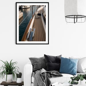May include: A framed wall art print featuring a high-angle view of a yacht deck with teak wood planks, a polished chrome cleat, and a coiled rope. The scene captures the side of the boat adjacent to dark blue harbour water and a stone pier.