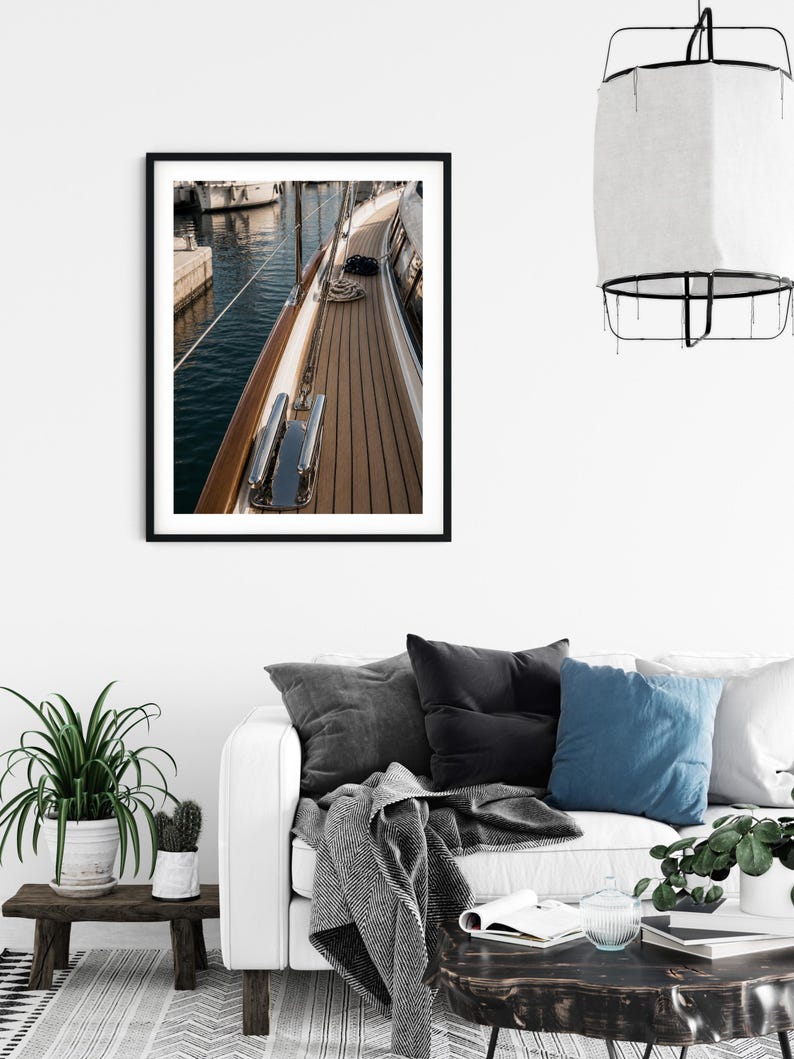 May include: A framed wall art print featuring a high-angle view of a yacht deck with teak wood planks, a polished chrome cleat, and a coiled rope. The scene captures the side of the boat adjacent to dark blue harbour water and a stone pier.