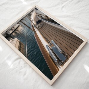 May include: A framed photograph showing a high-angle view of a wooden boat deck with a polished metal cleat in the foreground. The image captures the teak planks, the dark blue water reflecting the boat's side, and rigging lines extending upwards.