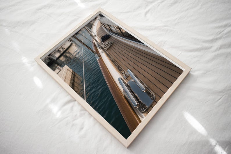 May include: A framed photograph showing a high-angle view of a wooden boat deck with a polished metal cleat in the foreground. The image captures the teak planks, the dark blue water reflecting the boat's side, and rigging lines extending upwards.