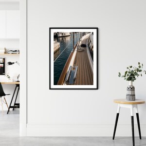 May include: A framed wall art print featuring a high-angle view of a yacht deck with teak wood planking, polished chrome hardware, and a stainless steel railing. The scene captures the side of the boat docked next to calm dark blue water.
