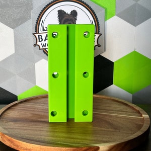 May include: Two lime green metal brackets with four holes each. The brackets are standing upright on a wooden surface.