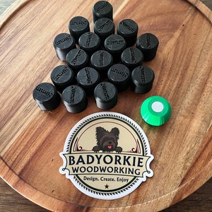 May include: A collection of 20mm black wooden plugs arranged in a triangle shape on a wooden surface. A green and white circular object is in the lower right corner of the image. A sticker with a dog and the text "BADYORKIE WOODWORKING Design. Create. Enjoy" is in the lower center of the image.