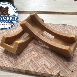 May include: Two brown wooden curved supports for a shelf or other woodworking project. The supports are made of wood and have a smooth finish. The supports are on a wooden surface with a herringbone pattern.