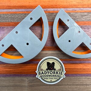 May include: Two gray, semi-circular woodworking jigs with orange accents and multiple holes. A sticker with a dog and the text "Badyorkie Woodworking" is also visible. The jigs are on a wooden surface with orange and brown stripes.