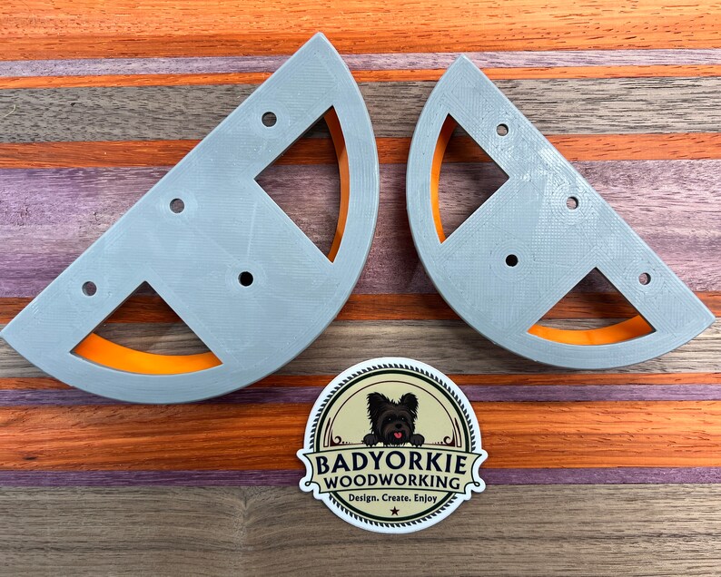 May include: Two gray, semi-circular woodworking jigs with orange accents and multiple holes. A sticker with a dog and the text "Badyorkie Woodworking" is also visible. The jigs are on a wooden surface with orange and brown stripes.