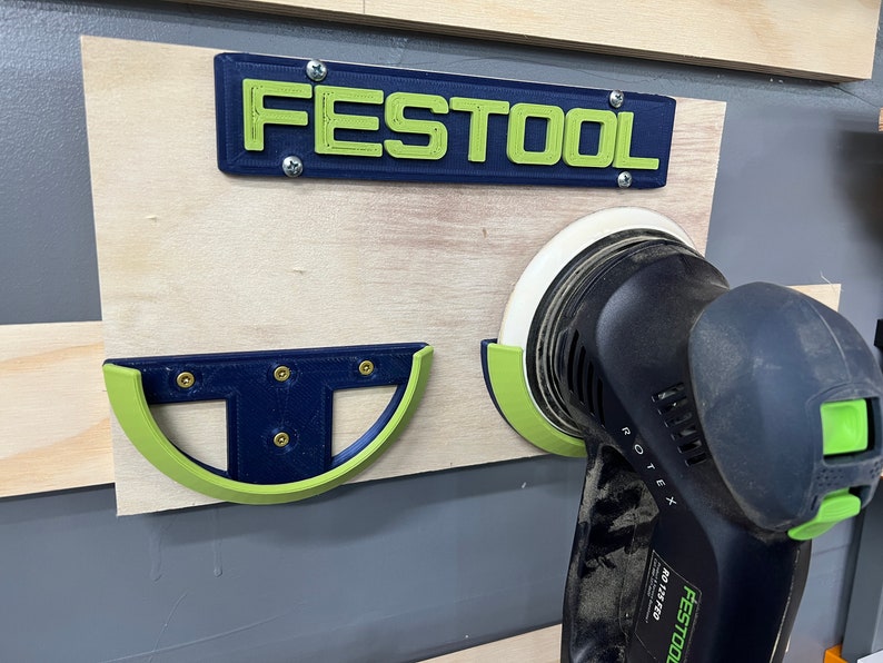 Pu&ograve; includere: Un organizer per utensili da parete con la scritta "FESTOOL" in lettere verdi su sfondo blu. Una levigatrice nera e verde &egrave; in un supporto. L'organizer &egrave; montato su una tavola di legno.