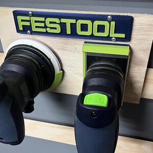 May include: A wooden workbench with a blue and green "FESTOOL" sign and two power sanders. The sign is mounted above the sanders. The sanders are black and gray with green accents. The workbench is made of light-colored wood.