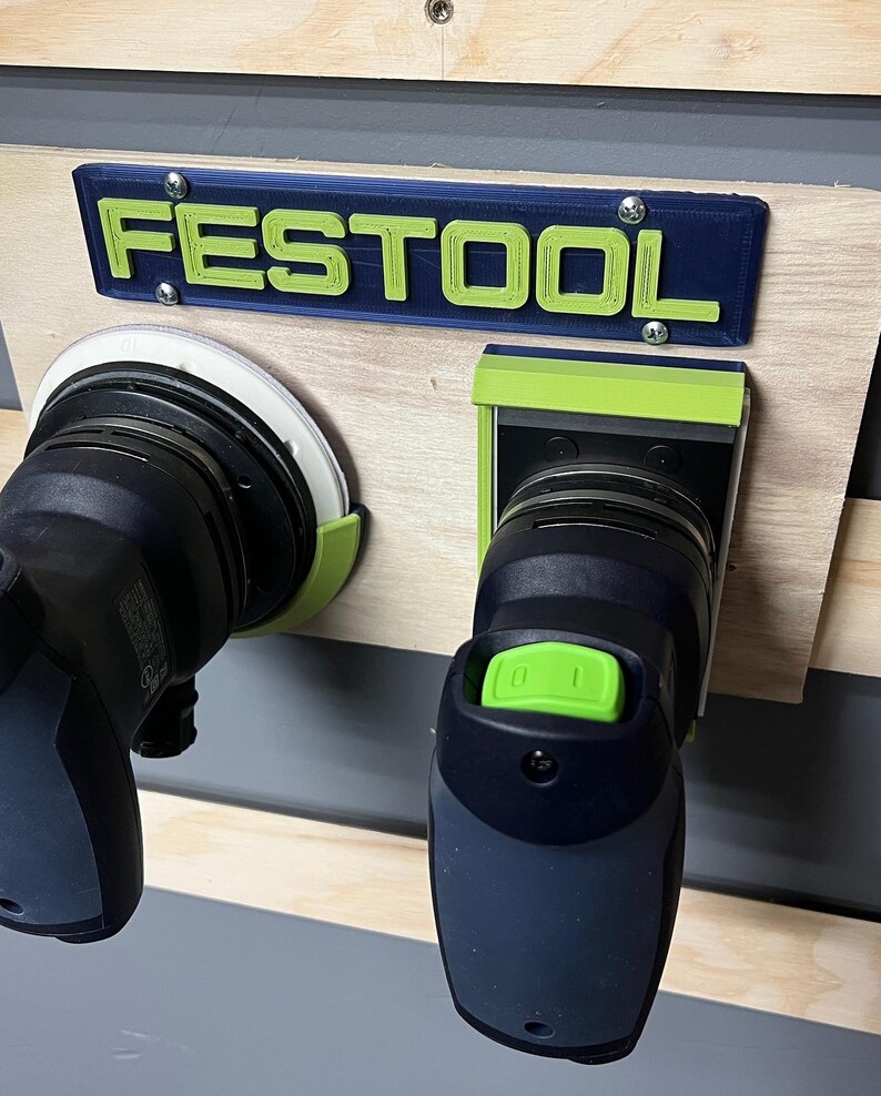 May include: A wooden workbench with a blue and green "FESTOOL" sign and two power sanders. The sign is mounted above the sanders. The sanders are black and gray with green accents. The workbench is made of light-colored wood.