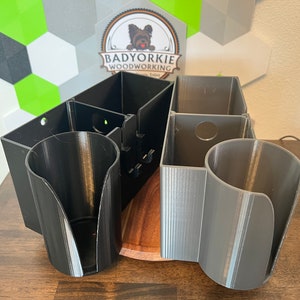 May include: A black and gray 3D printed wall mounted organizer with a wooden base. The organizer has multiple compartments and cup holders. The text "BADYORKIE WOODWORKING" is visible on the top of the organizer.