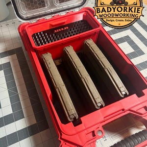 May include: A red storage case with a clear lid and a black handle. Inside are three brown magazines and a compartment labeled ".223/5.56". The case has a logo that says "Badyorkie Woodworking".