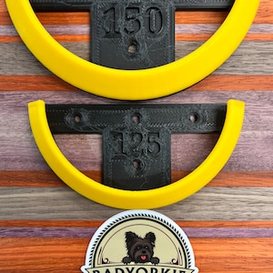 May include: Two black and yellow wooden wall mounted coat hooks with the numbers 150 and 125 printed on them. The hooks are on a wooden background with a sticker of a dog with the text "Badyorkie Woodworking Design. Create. Enjoy."