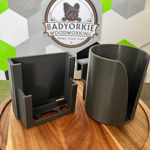 May include: A gray 3D printed phone stand and cup holder. The stand has a slot for a phone and a small shelf for charging cables. The cup holder is tall and cylindrical. Both items are sitting on a wooden surface.