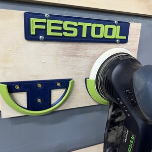 May include: A wall-mounted tool organizer with the word "FESTOOL" in green letters on a blue background. A black and green sander is in a holder. The organizer is mounted on a wooden board.