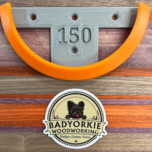 May include: A gray, T-shaped bracket with the number "150" and an orange curved piece. A sticker with a black dog and the text "Badyorkie Woodworking" is at the bottom. The background is a wooden surface with orange, purple, and brown stripes.