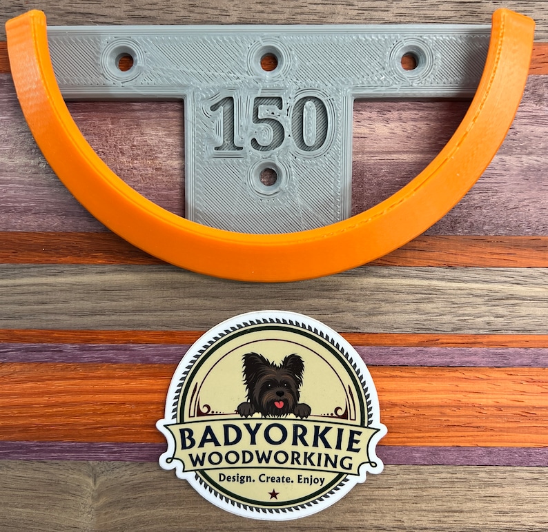 May include: A gray, T-shaped bracket with the number "150" and an orange curved piece. A sticker with a black dog and the text "Badyorkie Woodworking" is at the bottom. The background is a wooden surface with orange, purple, and brown stripes.