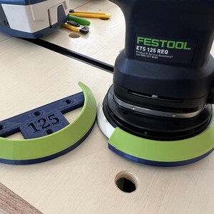 Pu&ograve; includere: Una levigatrice Festool ETS 125 REQ con un tampone di levigatura verde e blu. Una protezione per tampone di levigatura verde e blu abbinata con il numero "125" &egrave; accanto alla levigatrice. La levigatrice &egrave; nera e verde.
