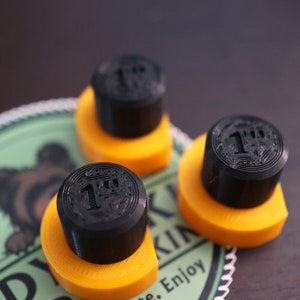 May include: Three black and orange 1-inch diameter stamps are displayed. Each stamp has a black cylindrical top with the number "1" printed on it, sitting on an orange base. The stamps are on a green and white background with a bear illustration.