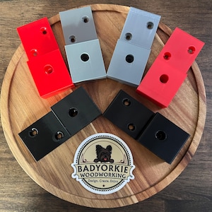 May include: An overhead shot displays several rectangular woodworking jigs in red, gray, and black, each with drilled holes. They are arranged on a wooden serving tray. A sticker with a dog and the text "Badyorkie Woodworking" is also visible.
