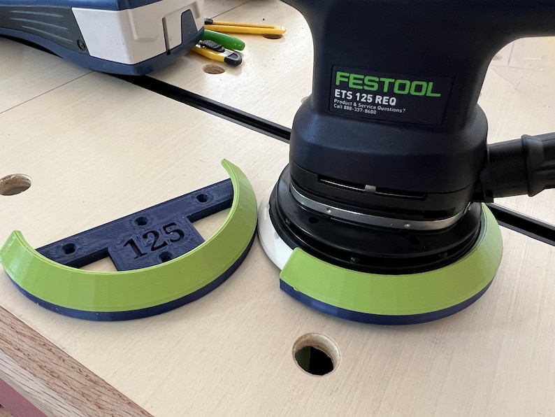 May include: A Festool ETS 125 REQ random orbital sander with a green and blue sanding pad. A matching green and blue sanding pad guard with the number "125" is next to the sander. The sander is black and green.
