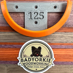 Pu&ograve; includere: Un supporto a parete grigio stampato in 3D con il numero "125" e un supporto curvo arancione. Sotto c'&egrave; un adesivo "Badyorkie Woodworking" con un cane. Il design include il testo "Design. Create. Enjoy."