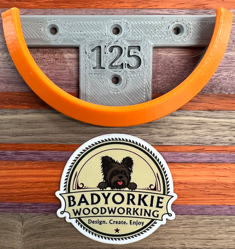 Pu&ograve; includere: Un supporto a parete grigio stampato in 3D con il numero "125" e un supporto curvo arancione. Sotto c'&egrave; un adesivo "Badyorkie Woodworking" con un cane. Il design include il testo "Design. Create. Enjoy."