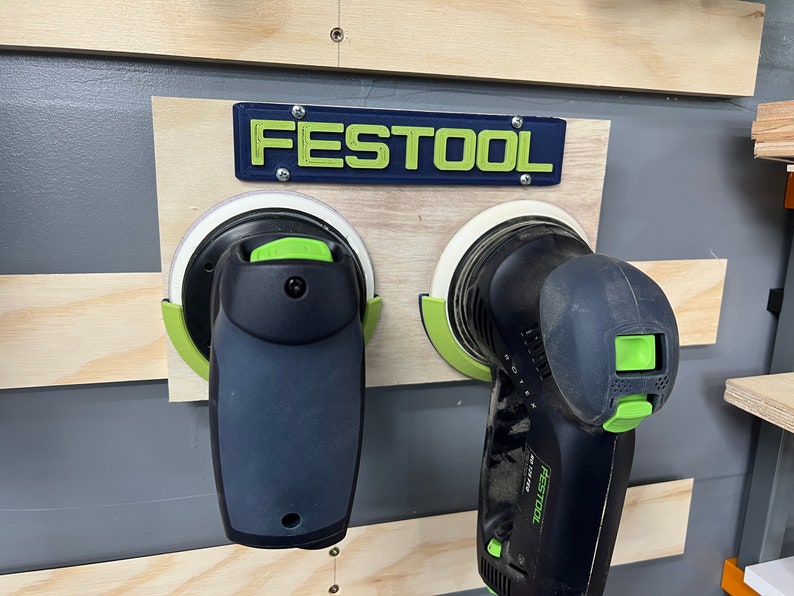 Pu&ograve; includere: Due levigatrici elettriche nere e verdi montate su una tavola di legno. Il marchio "FESTOOL" &egrave; visualizzato sopra le levigatrici in lettere verdi su sfondo blu. Le levigatrici hanno platorelli bianchi.