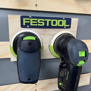 May include: Two black and green power sanders mounted on a wooden board. The brand name "FESTOOL" is displayed above the sanders in green letters on a blue background. The sanders are attached to a gray wall.