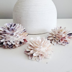 May include: Three decorative flower-shaped ornaments crafted from layered white and purple seashells. The shells are arranged in a dense, circular pattern to resemble blooming petals, with varying sizes of the floral structures displayed together.