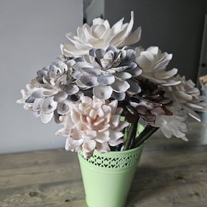May include: A bouquet of handcrafted wooden flowers in a light green metal pot. The flowers are various shades of white, grey, and brown. The pot has a decorative cut-out design. The arrangement sits on a wooden surface.