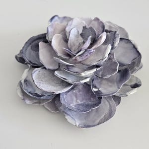 May include: A decorative flower-shaped object crafted from layered, overlapping seashell fragments. The shells are painted in varying shades of muted purple, lavender, and white, arranged in a circular, blooming pattern resembling a succulent plant.
