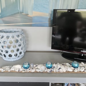 May include: A decorative display featuring a light blue, open-weave lantern and a wooden base adorned with seashells and three blue glass orbs. The display sits on a gray surface, with a painting and a television in the background.