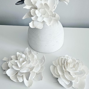 May include: A white, ribbed, rounded vase containing several white, layered, petal-like floral decorations, with two additional matching white floral pieces resting separately in front of the vase on a flat surface.