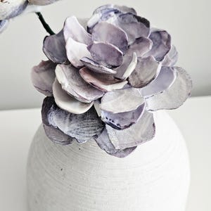 May include: A decorative flower sculpture crafted from layered purple and white seashells, arranged in a bloom shape and attached to a thin green stem, displayed inside a round, textured white ceramic vase with horizontal ridges.