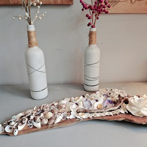 May include: A driftwood wall decor piece with a variety of seashells and a white flower. Two white painted wine bottles with twine accents are in the background.