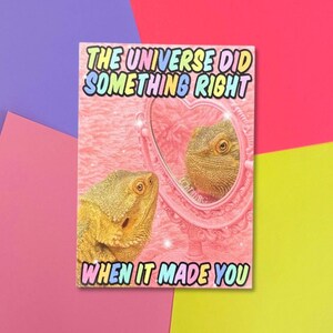 Könnte beinhalten: Eine rechteckige Grußkarte mit einer Bartagame, die in einen rosa herzförmigen Spiegel schaut. Der Text lautet THE UNIVERSE DID SOMETHING RIGHT WHEN IT MADE YOU in bunten Buchstaben. Der Hintergrund ist rosa strukturiert mit funkelnden Lichteffekten.