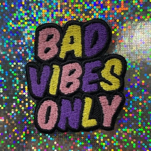 Könnte beinhalten: Gestickter Aufnäher mit dem Text "BAD VIBES ONLY" in einer Blasenschrift. Die Buchstaben sind schwarz umrandet und mit rosa, gelb und lila gefüllt.