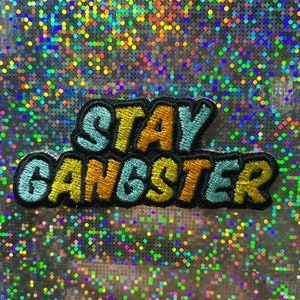 Könnte beinhalten: Ein gestickter Aufnäher mit dem Text "Stay Gangster" in Blau, Gelb und Schwarz.
