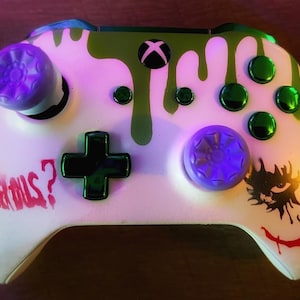 Könnte beinhalten: Ein weißer Xbox-Controller mit grünen und violetten Akzenten. Der Controller hat ein Joker-Thema mit den Worten "Why so serious?" auf der Seite. Der Controller hat auch ein grünes Schleim-Design auf der Oberseite.