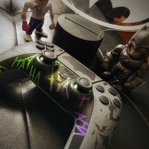 May include: A black and gray video game controller with green and purple accents. The controller has a design of a Joker character with the words "HA HA" printed on it.