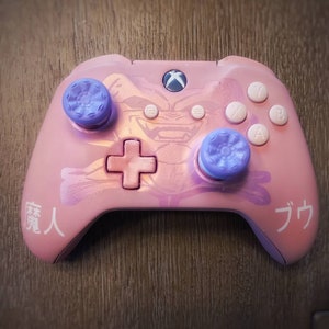 Op de afbeelding: Een roze Xbox-controller met een paars en blauw ontwerp. De controller heeft een schedel en gekruiste beenderen ontwerp aan de voorkant en de woorden "Demon" en "Goku" in het Japans geschreven aan de zijkanten.
