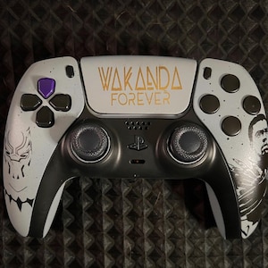 Puede incluir: Un mando de PlayStation 5 blanco y negro con un diseño personalizado que presenta una pantera negra y las palabras "Wakanda Forever".