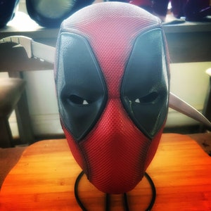 Op de afbeelding: Een rood en zwart Deadpool-masker met een textuur. Het masker staat op een zwarte standaard en heeft een scherp, zilveren mes erachter.