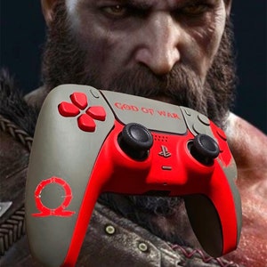 May include: A gray and red PlayStation 5 DualSense controller with a custom God of War design. The controller features a red and gray color scheme with a gray and red symbol on the left side. The controller has the words "God of War" printed on the top.