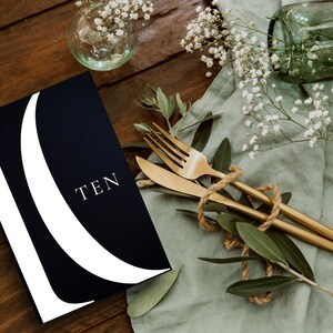 May include: A black and white table number card with the word "TEN" printed in white on a black background. The card is on a wooden table with a green cloth and gold cutlery.