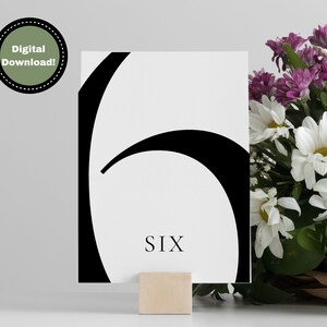 Puede incluir: Tarjeta de n&uacute;mero de mesa en blanco y negro con el n&uacute;mero seis y la palabra "SIX" impresa en negro. La tarjeta est&aacute; en un soporte de madera.