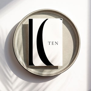 Puede incluir: Una tarjeta blanca con una letra C estilizada en negro y la palabra "TEN" impresa en negro. La tarjeta est&aacute; sobre un plato gris.