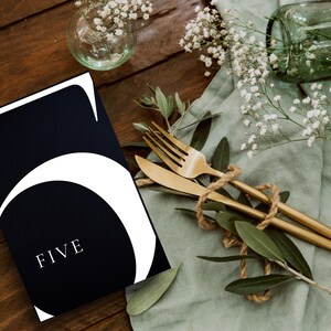 May include: A black and white table number card with the number "5" and the word "FIVE" in white text. The card is on a wooden table with a green cloth, gold cutlery, and white flowers.
